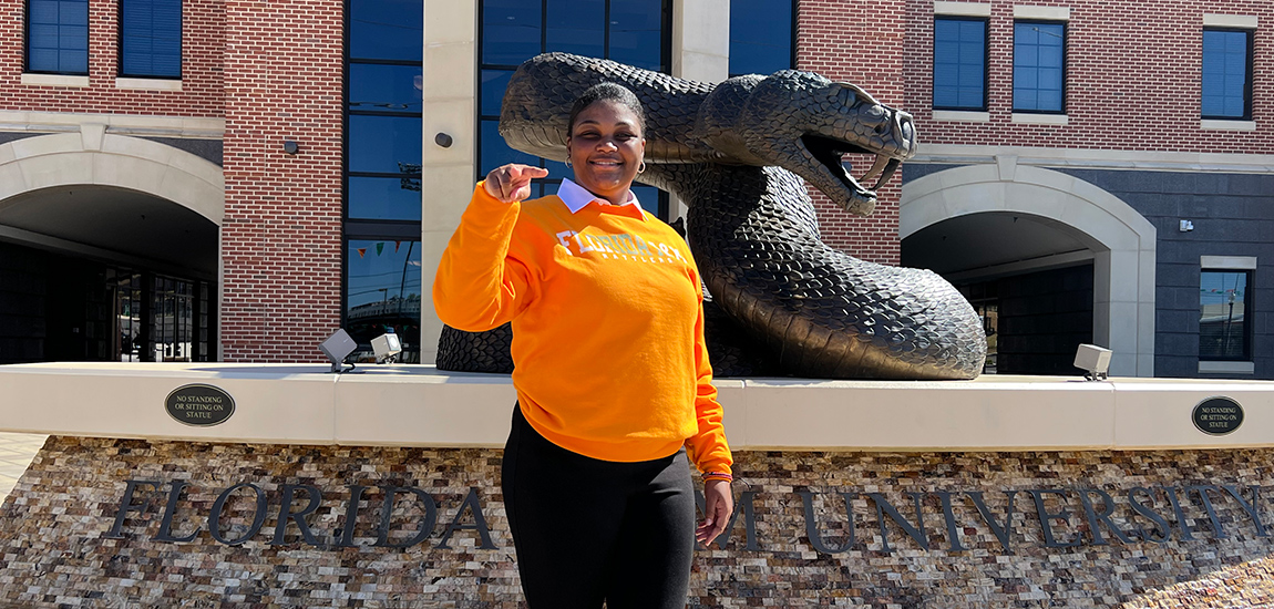 A young woman smiling at the camera, she has on an orange sweatshirt and black pants, she stands in front of a snake statue